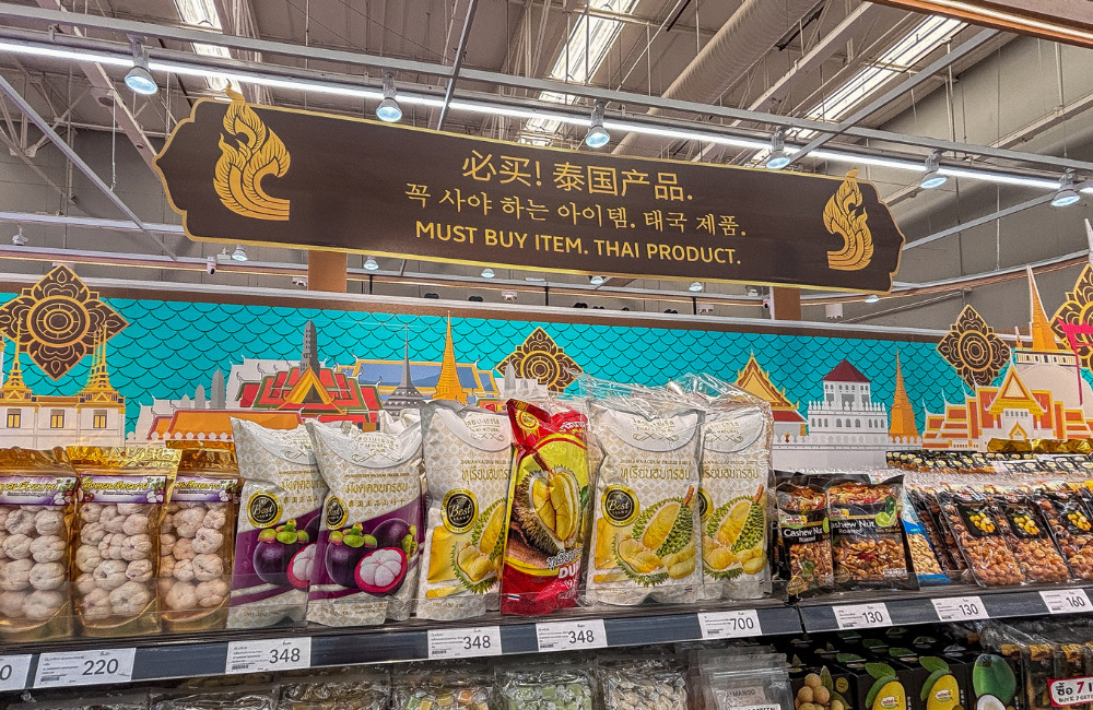 Display of Thai products at Lotus's Sukhumvit 50 featuring snacks like durian chips, mangosteen crisps, and cashew nuts with a decorative Thai-themed background.