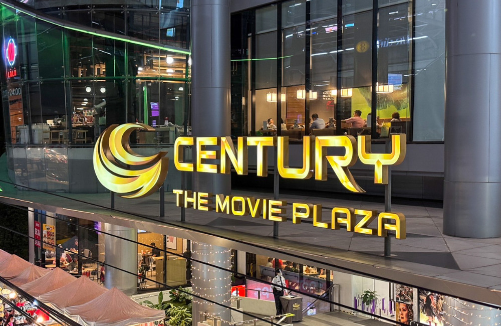 Exterior view of Century The Movie Plaza Sukhumvit at night, showcasing its illuminated golden signage and bustling activity inside and outside the plaza.