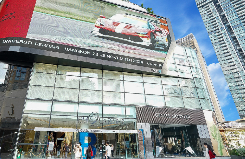 Front view of EmQuartier mall in Bangkok, showcasing the Glass Quartier entrance with a large digital screen displaying a Ferrari advertisement. The façade includes luxury brand stores like Gentle Monster and Zara, with modern skyscrapers in the background.