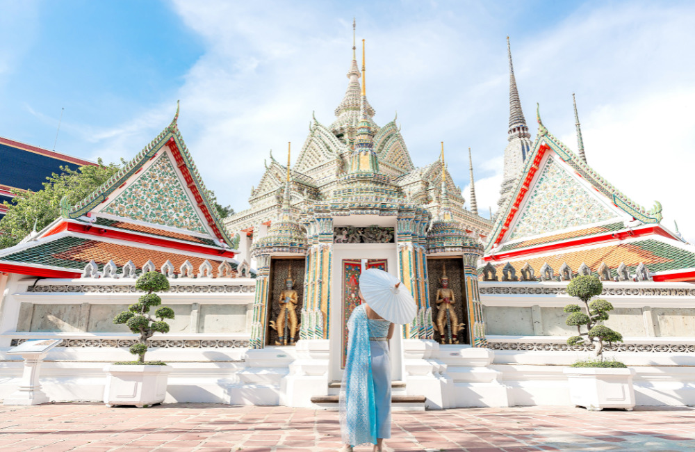 A visitor in an elegant traditional Thai dress holding a white parasol, standing before a magnificent temple in Bangkok during the Songkran 2026 festival.