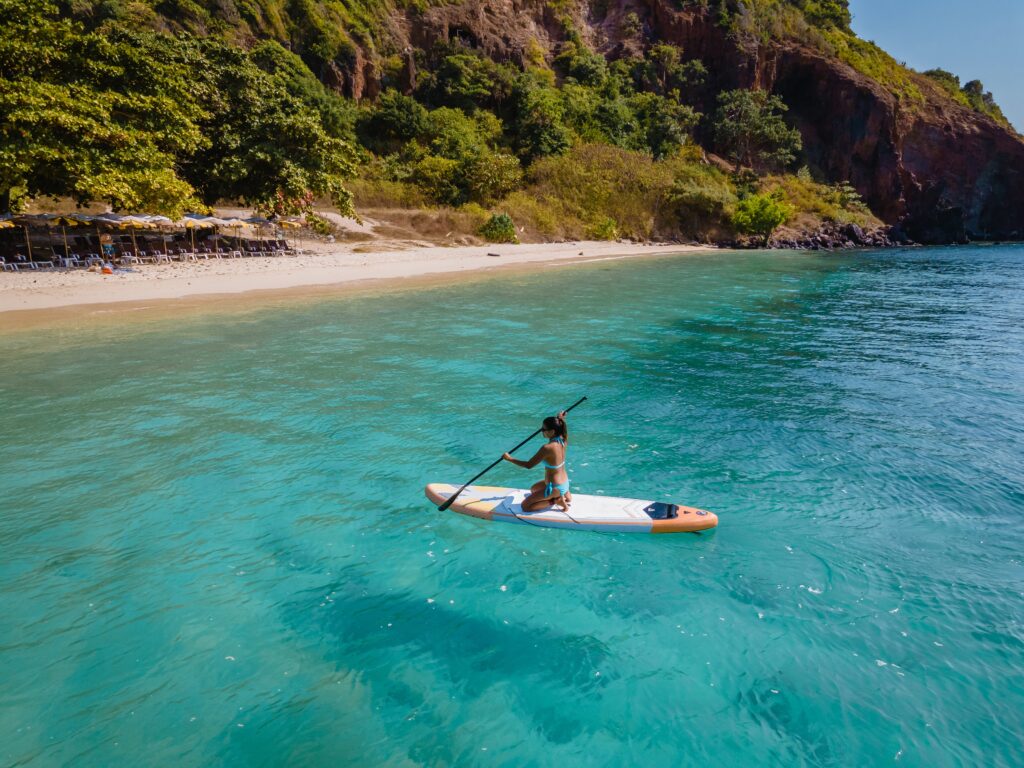 A woman in blue swimwear paddles a stand-up paddleboard across clear turquoise water beside a quiet, tree-lined beach on Koh Larn.
