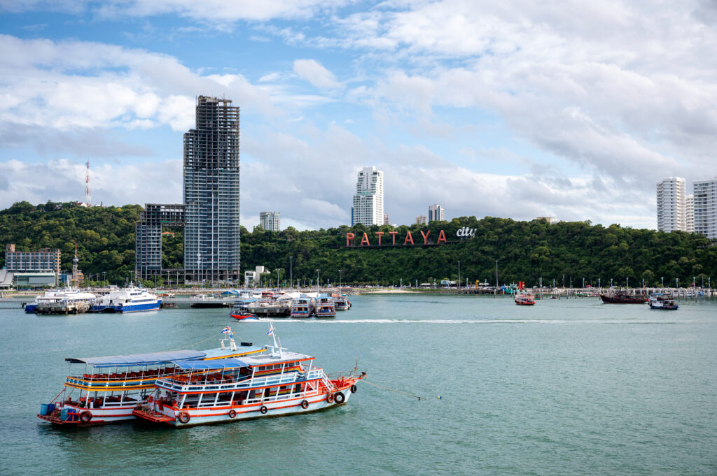 A colorful passenger ferry crosses Pattaya Bay near Bali Hai Pier, backed by green hills, high-rise buildings, and the Pattaya City sign.