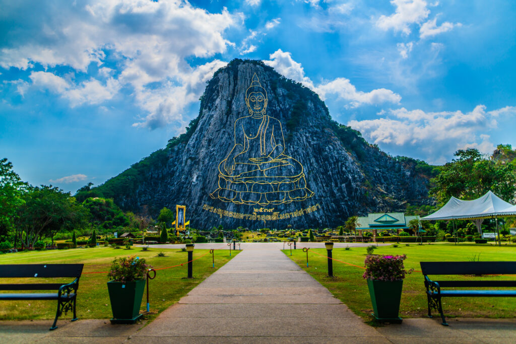 วิวหน้าทางเดินเข้าสู่เขาชีจรรย์ พัทยา พร้อมพระพุทธรูปแกะสลักทองคำบนหน้าผา ท่ามกลางสวนเขียวขจีและท้องฟ้าโปร่ง