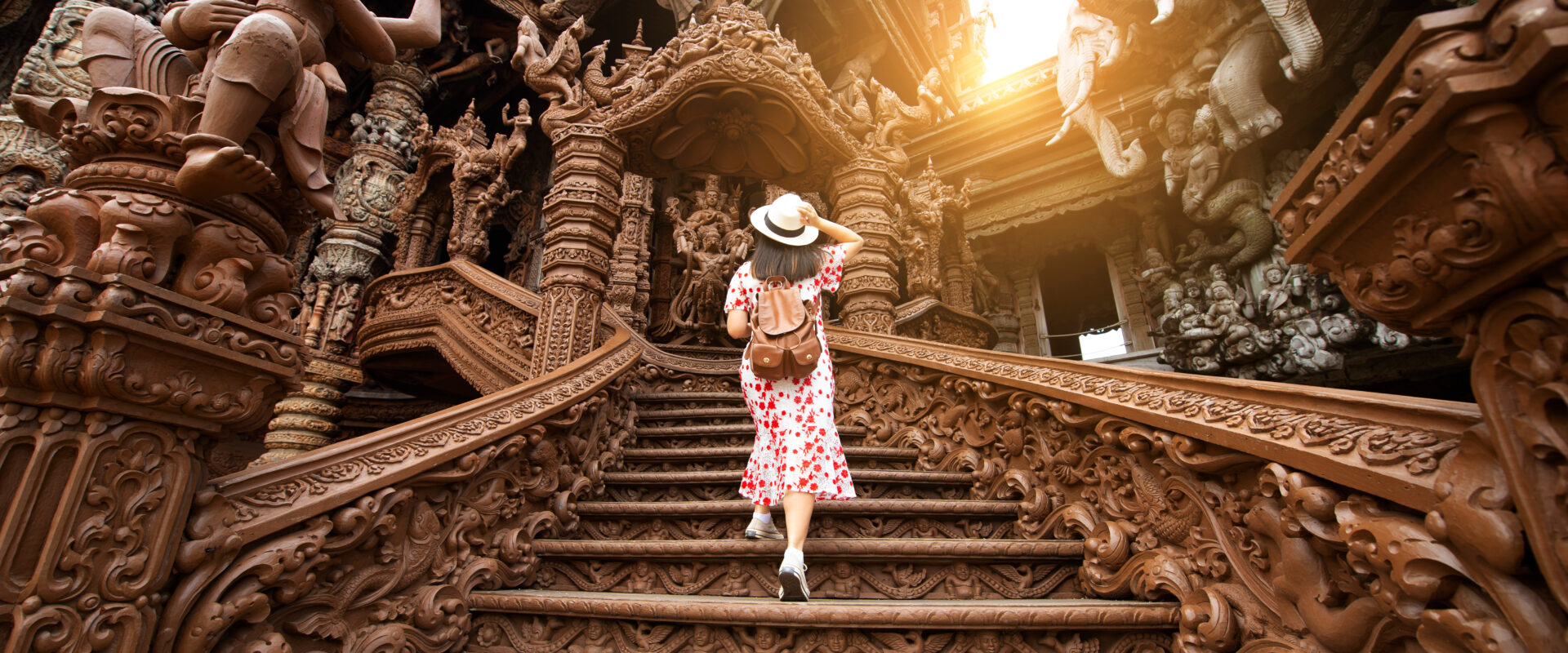 A woman in a red and white dress and sun hat walks up the intricately carved wooden staircase of the Sanctuary of Truth in Pattaya, Thailand. The sun shines through the structure, highlighting the detailed sculptures and ancient Thai craftsmanship.