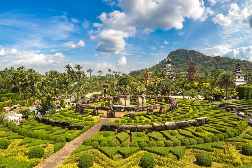 วิวสวนหินจำลองและพุ่มไม้ตัดแต่งเป็นวงกลมอย่างวิจิตรที่สวนนงนุช พัทยา โดยมีฉากหลังเป็นภูเขาและเจดีย์สไตล์เอเชียกลางธรรมชาติ