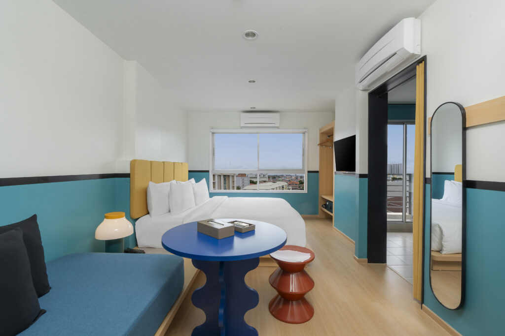 A modern and bright New Deluxe King Room with a city view, featuring a comfortable king-sized bed, a stylish blue accent table, a cozy lounge area, and a wall-mounted flat-screen TV. The room's design includes vibrant colors and contemporary decor, offering a welcoming atmosphere.