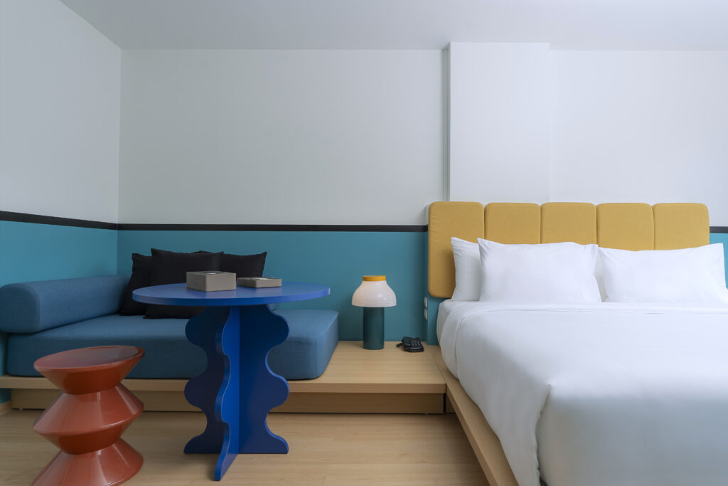 A modern Deluxe King Room with City View, featuring a comfortable king-sized bed with white linens, a yellow headboard, a cozy blue sofa, a stylish blue table, and a unique orange side table, offering a contemporary and minimalist design.