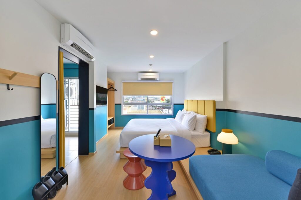 Colorful Pattaya hotel room with a double bed, blue sofa, round table, full-length mirror, balcony entrance, and a large window overlooking the city.