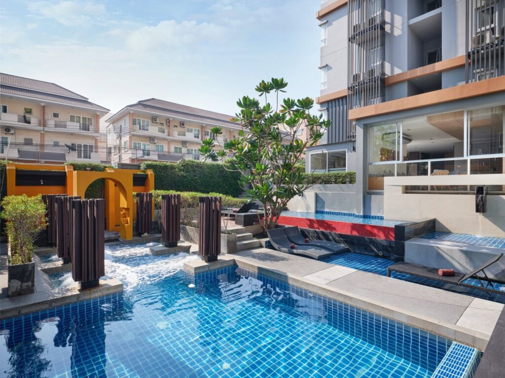 Outdoor swimming pool at a Pattaya hotel with blue-tiled water, built-in loungers, decorative fountains, greenery, and a modern guest building.